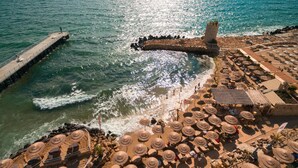An aerial view of a coastal area with a pier extending into the sea, a stone tower, and a beach with numerous sun umbrellas and lounge chairs. - Villa Chinka by Astor Garden Hotel (Varna)