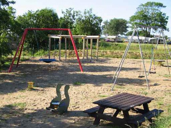 Un parque infantil con columpios y una mesa de picnic en una zona arenosa, con árboles al fondo.