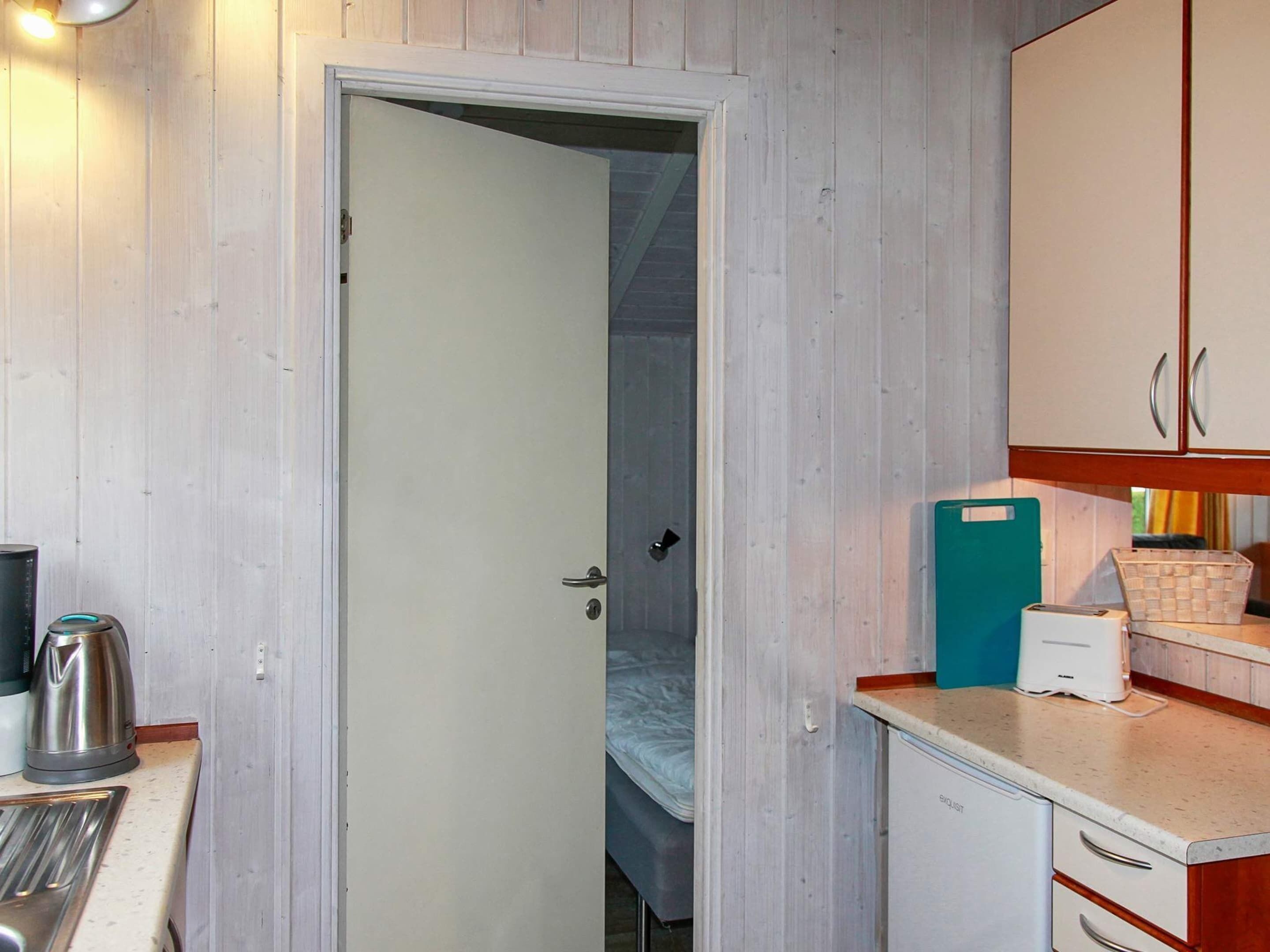 A compact kitchen with a white door, a bed in the adjacent room, a kettle, a toaster, and a small refrigerator.