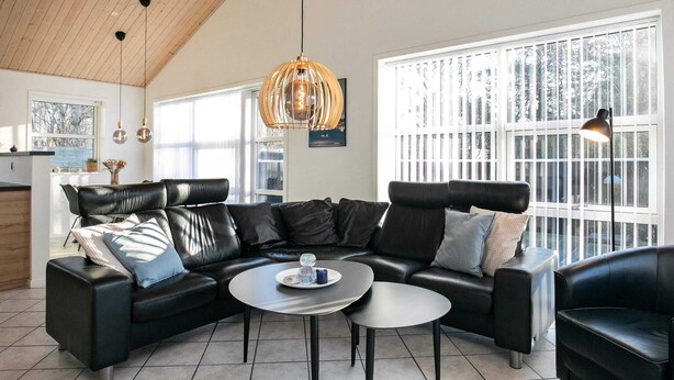 A modern living room with a black leather sectional sofa, a round coffee table, and a large window with blinds. - Splendid Holiday Home in Laeso near Museum Court (Læsø)