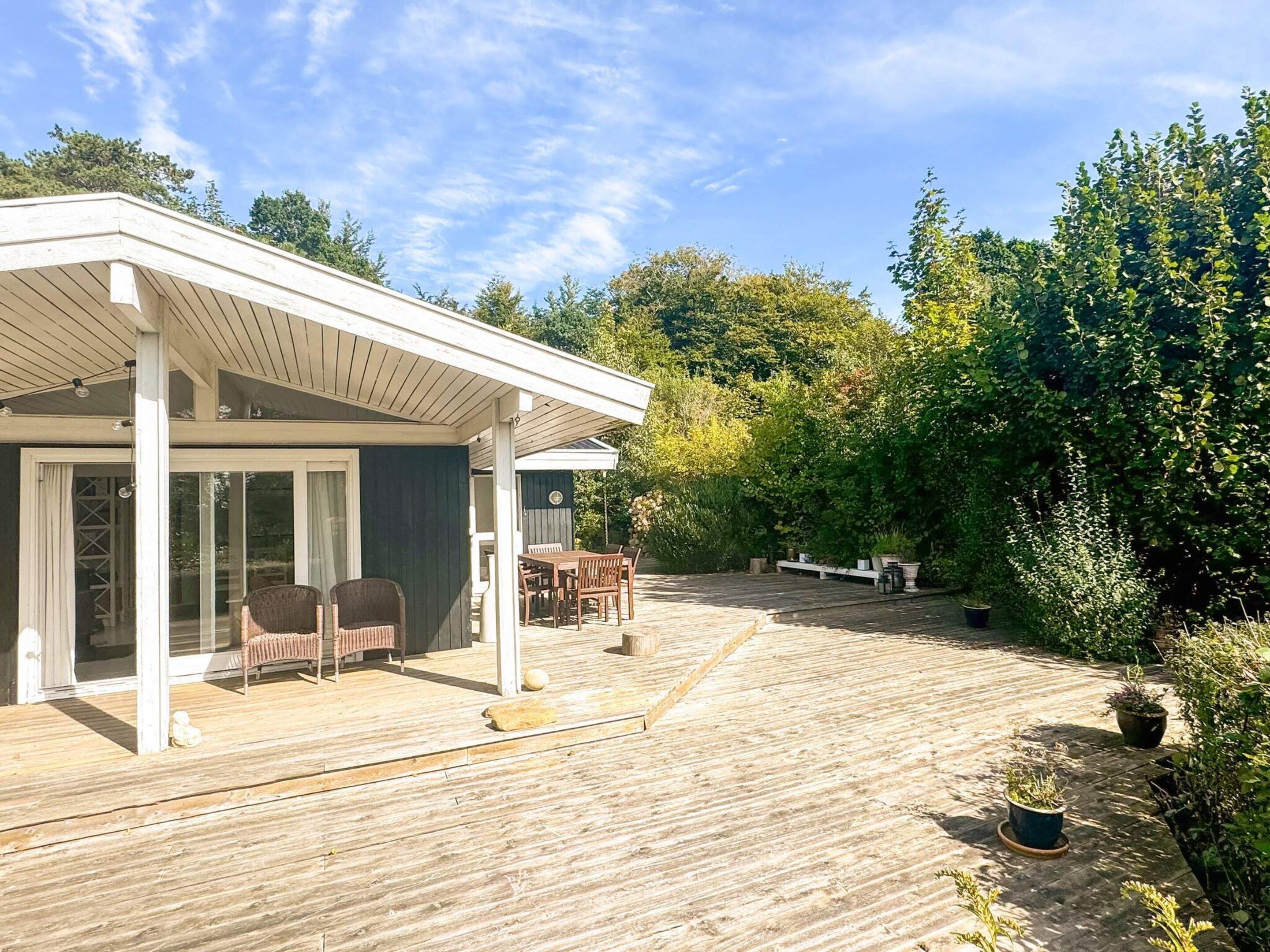 A covered outdoor area with a wooden deck, a seating area with chairs and a table, and a view of a garden with various plants and trees.