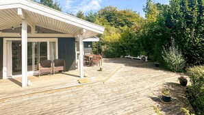 Uma área externa coberta com deck de madeira, um espaço de estar e vista para um jardim com diversas plantas.