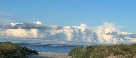 A sandy beach path leading to the sea with grassy dunes on either side and a clear sky with clouds.