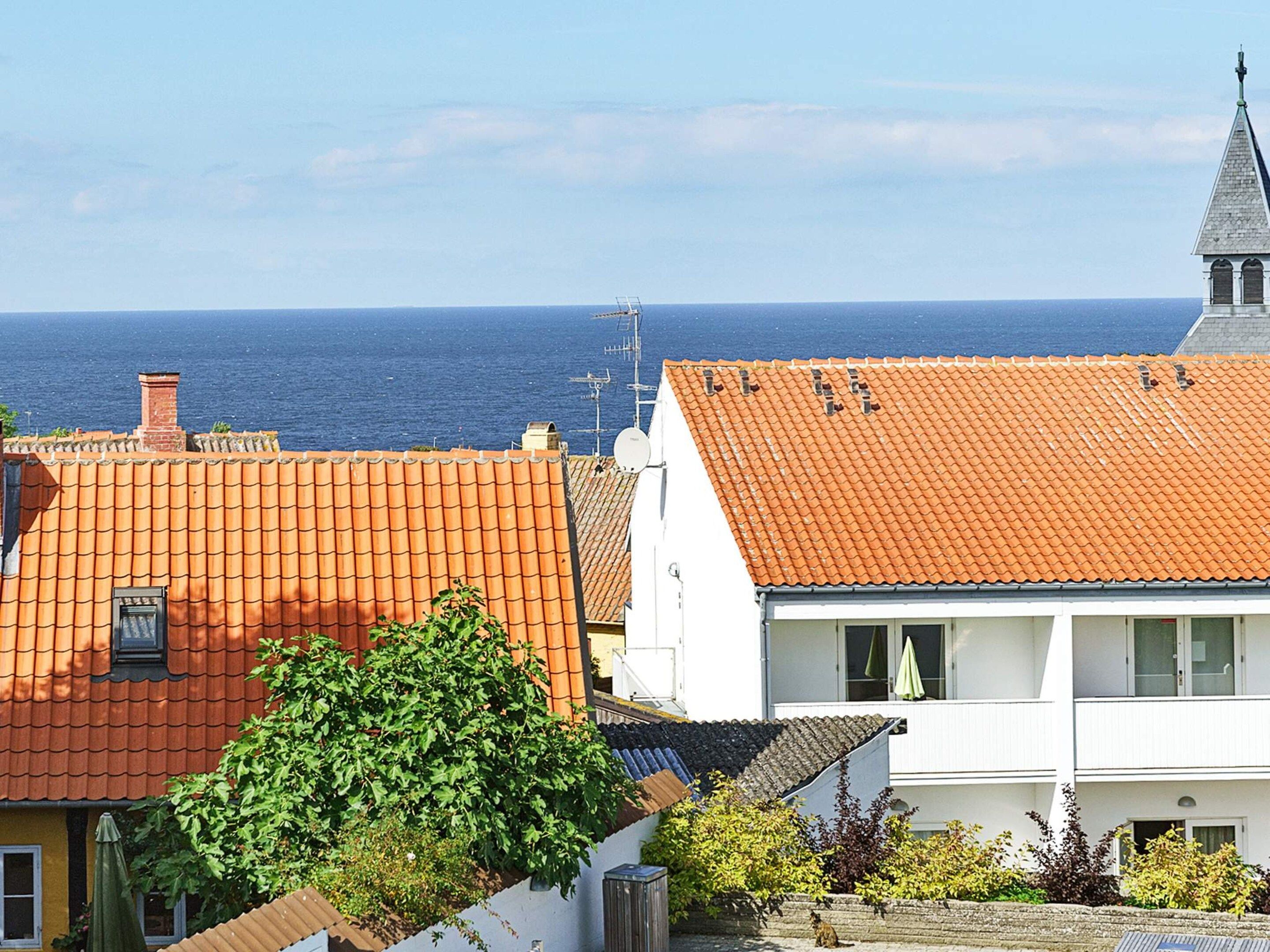 A coastal residential area with terracotta-tiled roofs, a white building with a balcony, and a clear view of the sea.
