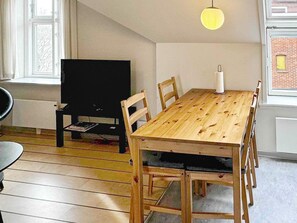 A dining area with a wooden table, chairs, a black television, and a window with curtains. - 4 Person Holiday Home in Gudhjem (Gudhjem)