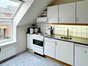 Cocina compacta con estufa, horno y lavavajillas integrados, armarios blancos y ventana que proporciona luz natural.