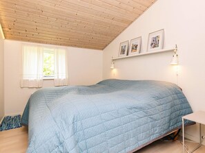 A bedroom with a bed, a window with curtains, a wall-mounted shelf with pictures, and a bedside table with a lamp.