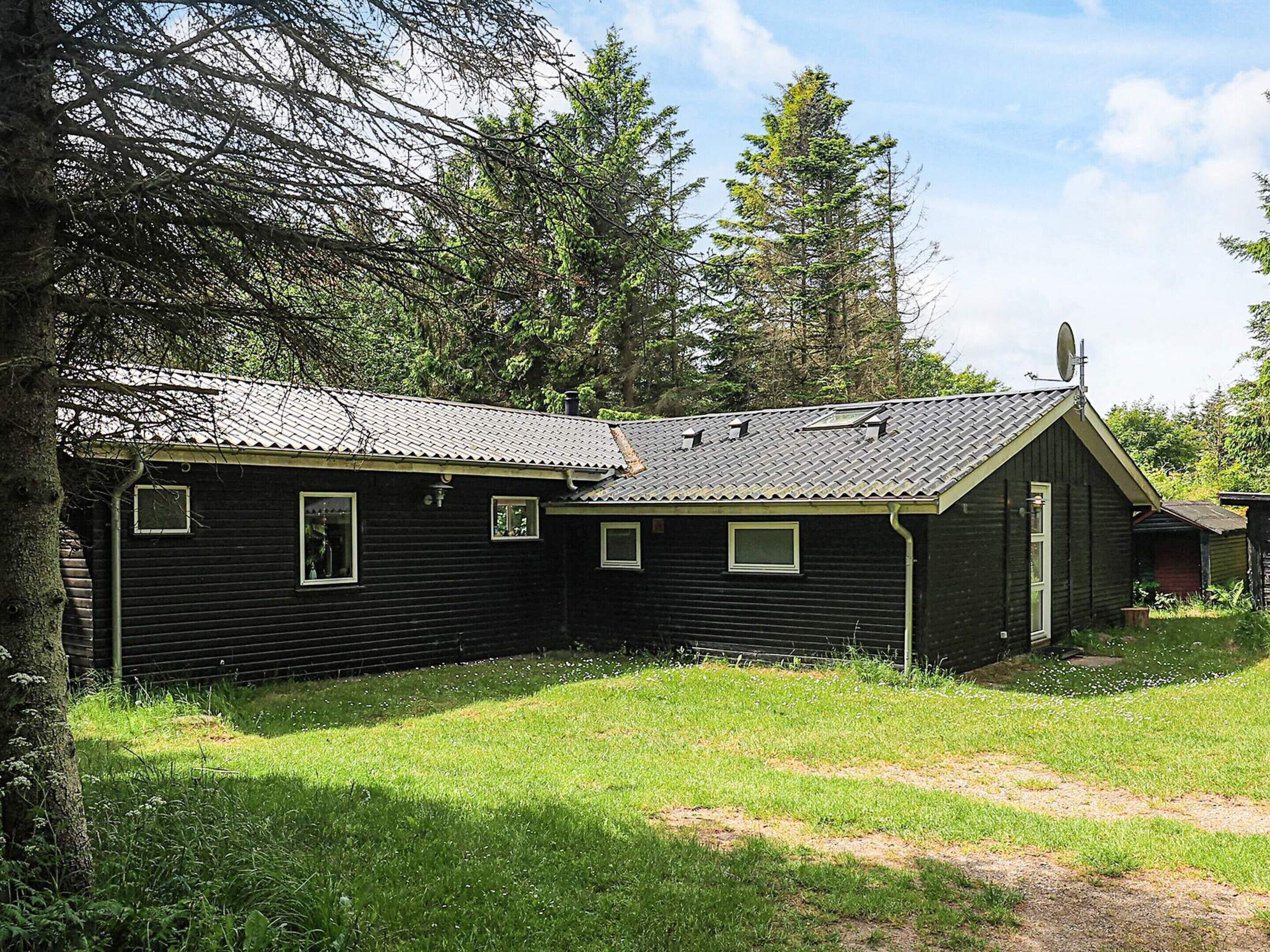 A small, single-story wooden cabin with a sloped roof, surrounded by a grassy area and dense trees.