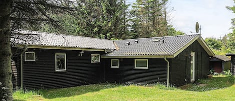 A small, single-story wooden cabin with a sloped roof, surrounded by a grassy area and dense trees.