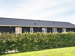 A long, dark-colored building with a tiled roof, white-framed windows, and a well-maintained hedge in front.