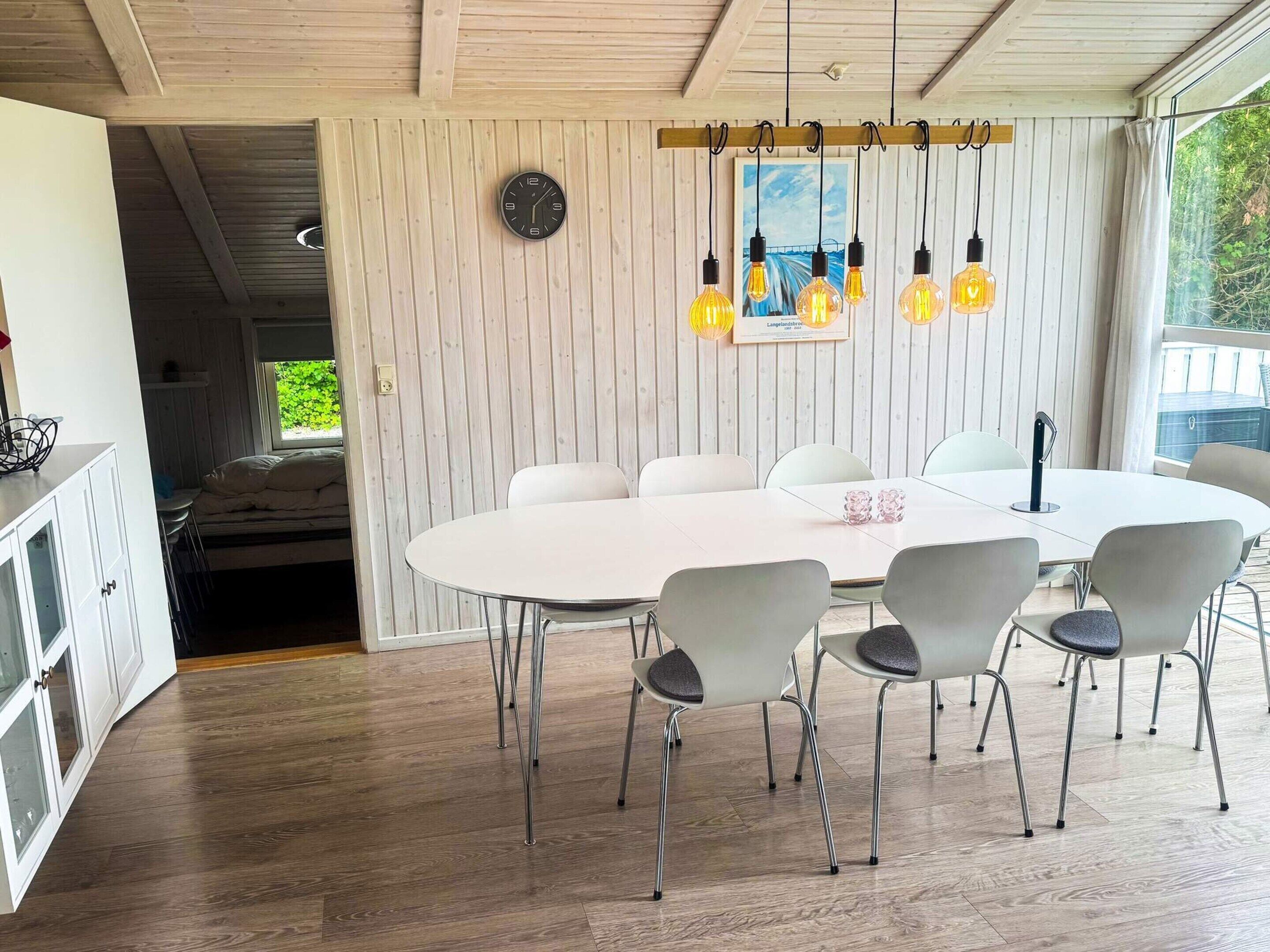 A modern dining area with a white table and chairs, a clock on the wall, and hanging light bulbs.