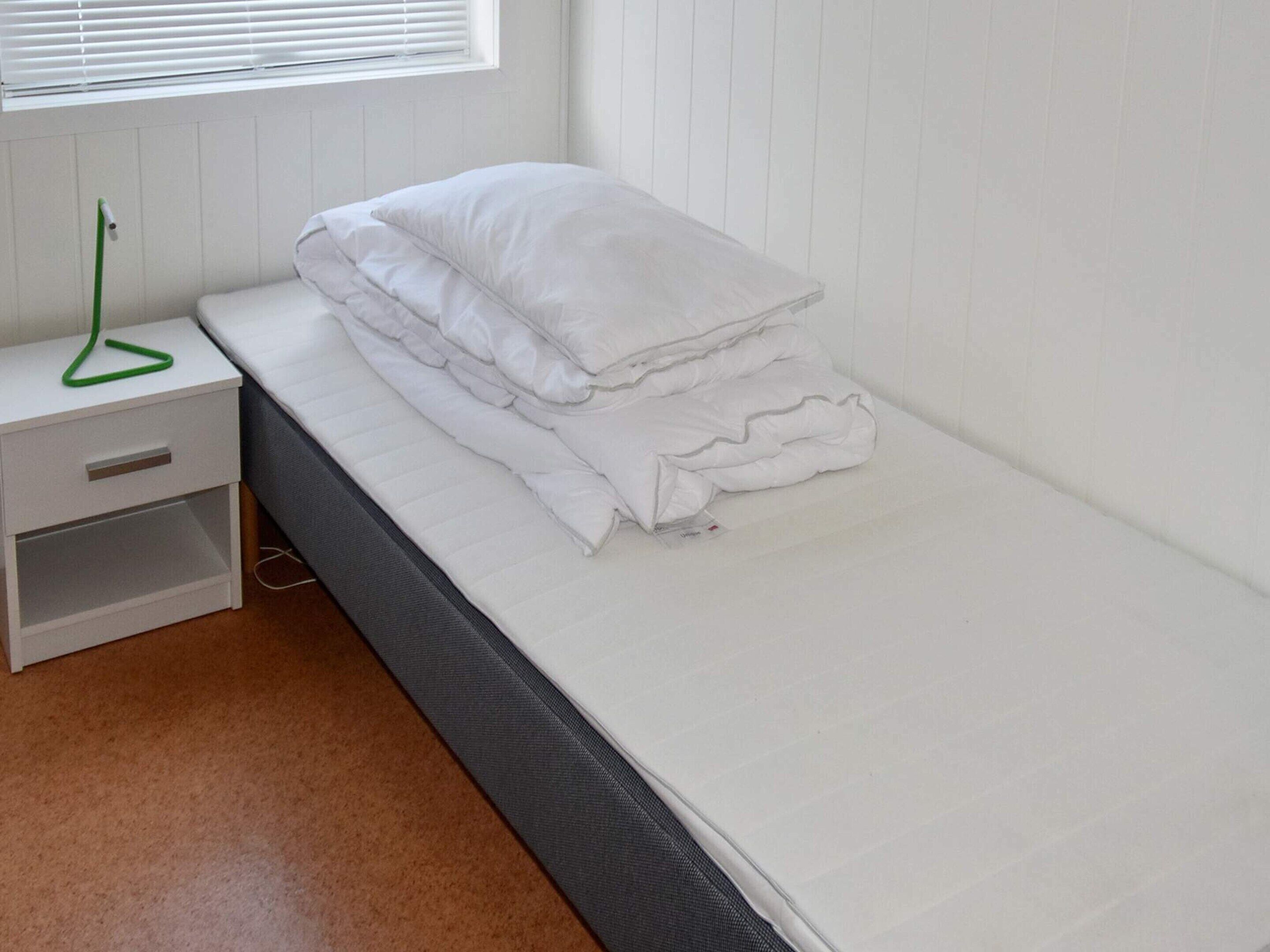 A single bed with white bedding, a white nightstand with a green object on it, and a window with blinds.