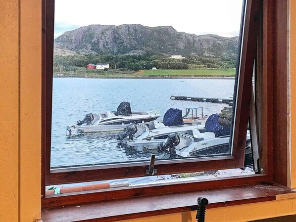 Vista desde una ventana: un muelle con barcos, una montaña al fondo y un edificio rojo.