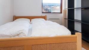 Quarto com cama de madeira, roupa de cama branca, uma estante preta e janela com vista para a cidade.