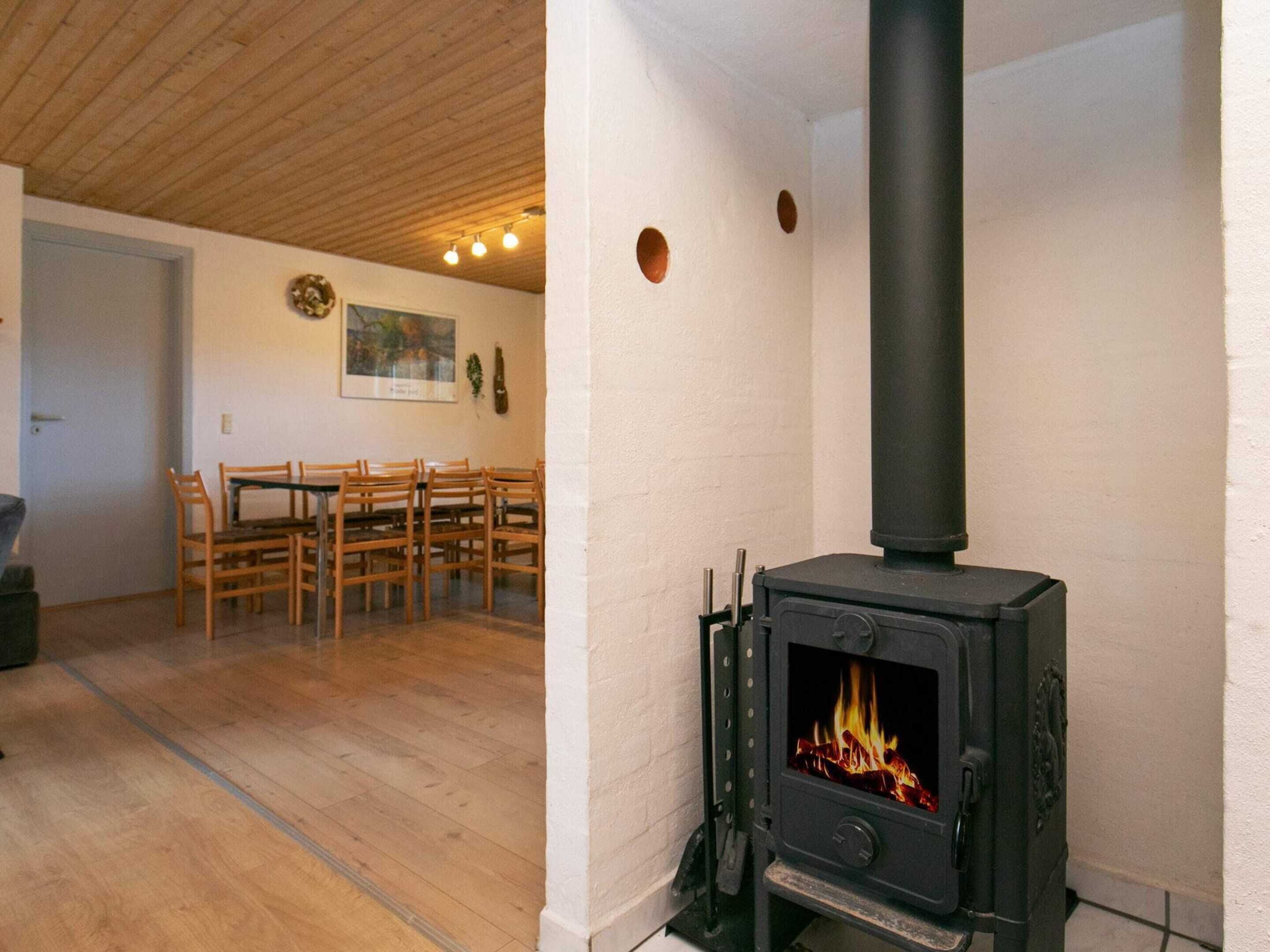 A room with a black wood-burning stove, wooden chairs, a dining table, and a white wall with a painting.