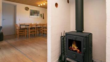 A room with a black wood-burning stove, wooden chairs, a dining table, and a white wall with a painting.