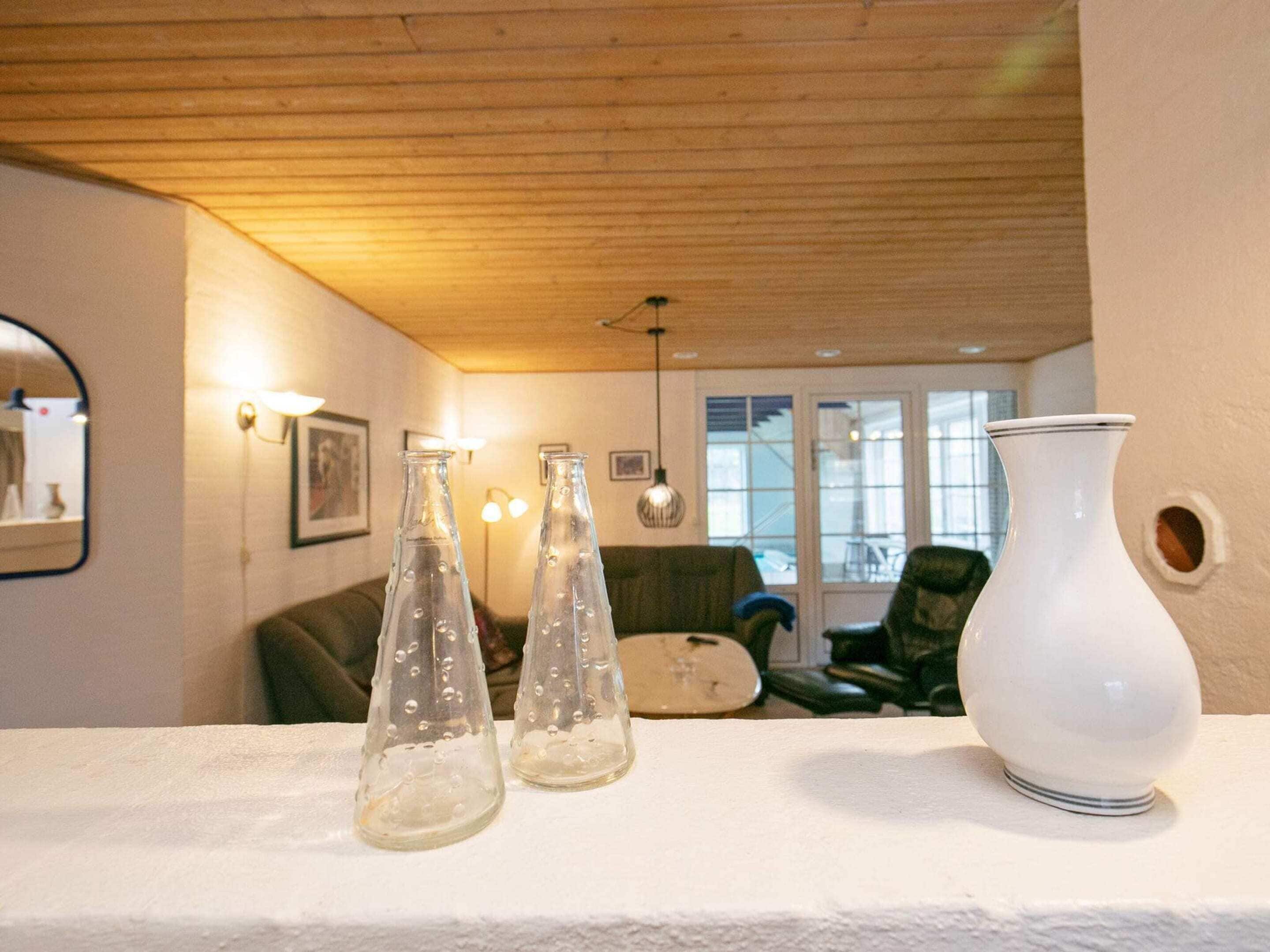 Una stanza con soffitto in legno, parete bianca e una finestra. Tre bottiglie di vetro su una superficie bianca e un vaso bianco.