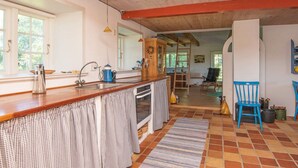 Uma cozinha com bancada de madeira, pia e fogão. Há uma janela com cortinas, uma cadeira e um tapete no chão.