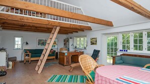 Um quarto amplo com mezanino, vigas de madeira, escrivaninha, cadeira, sofá e mesa de jantar com toalha xadrez.