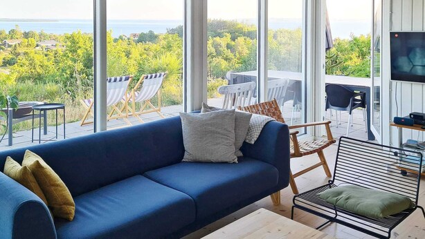 Un salon moderne avec un canapé bleu, une table basse en bois et une vue sur une forêt et un plan d’eau.