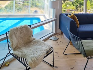 A room with a view of a swimming pool, a blue sofa with cushions, a fluffy chair, and a metal-framed chair.