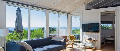 A spacious living area with a sofa, coffee table, and a TV. Large windows offer a view of the outdoors.