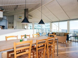 A spacious dining area with a wooden table and chairs, a kitchen in the background, and large windows offering views.