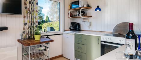 A compact kitchen with a built-in microwave, a stove, and a sink. There's a window with curtains, a small shelf with a lamp, and a bottle of wine on the counter.