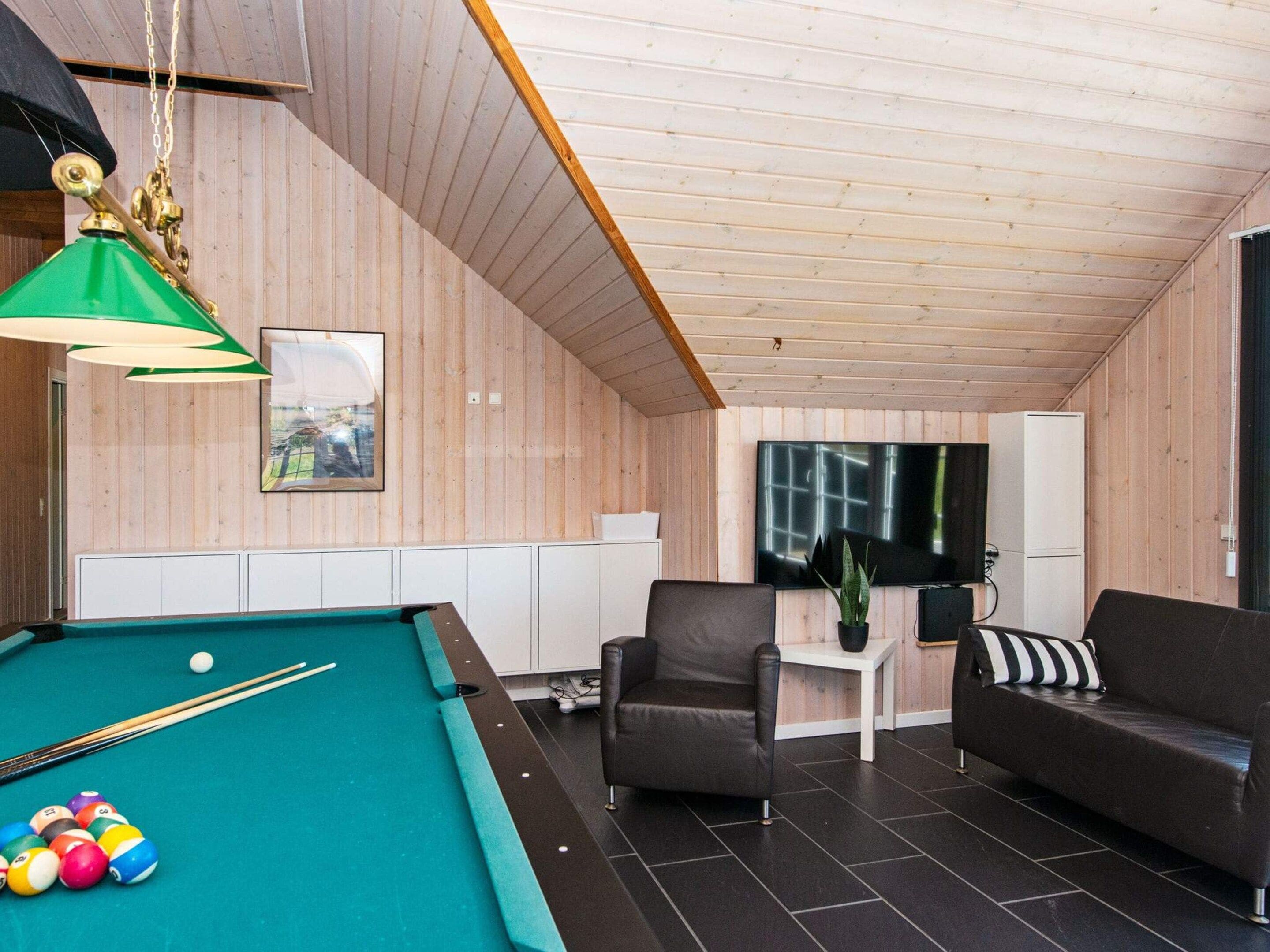 A pool table with balls, a sofa, a chair, a small table with a plant, and a television on the wall.