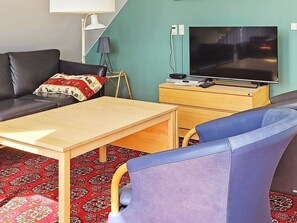 Un salon comprenant un canapé, une table basse en bois, un fauteuil bleu, un téléviseur posé sur un meuble en bois et un tapis à motifs.