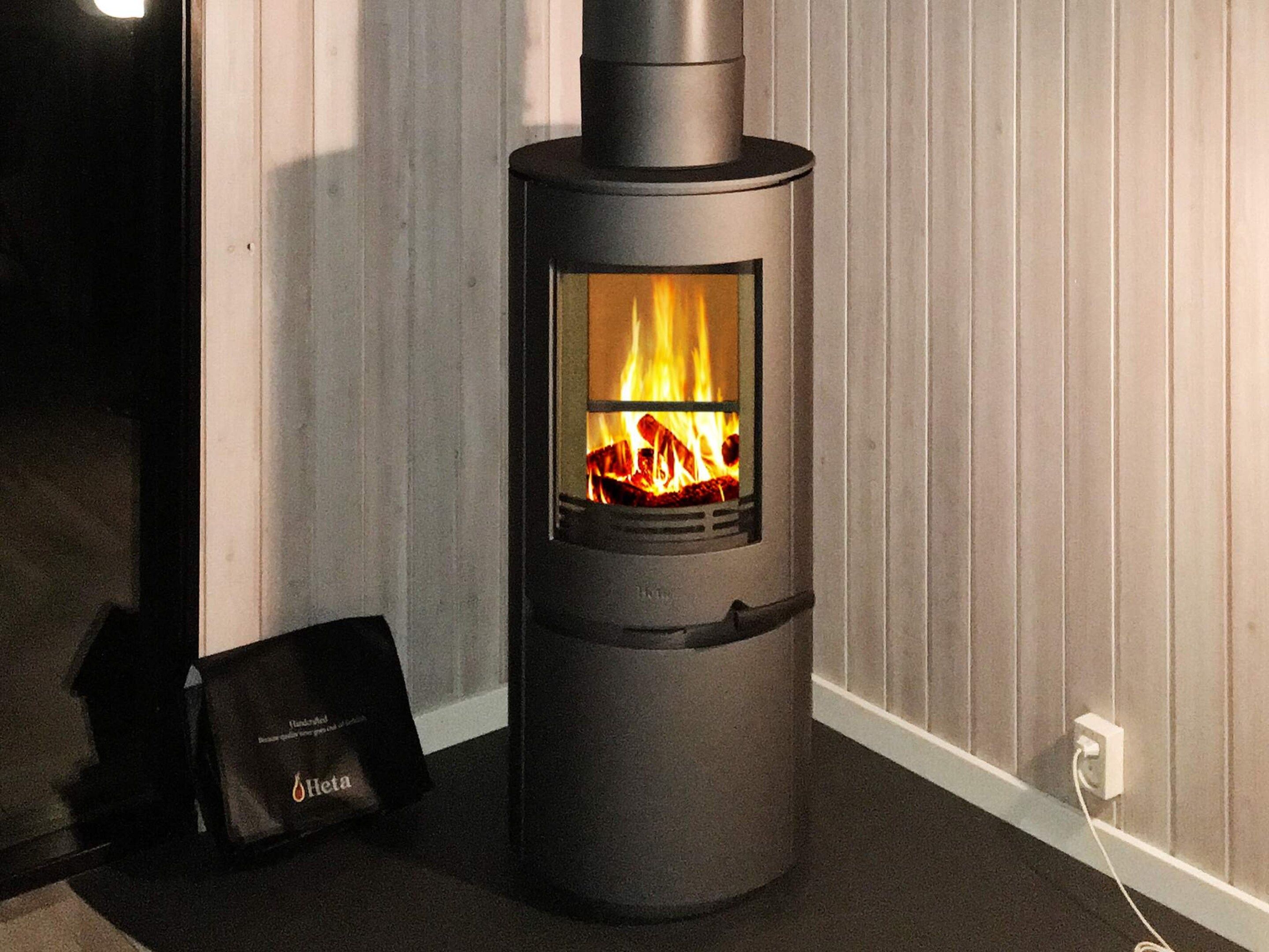 A modern wood-burning stove with a glass front, a black bag labeled 'Heta' beside it, and a power outlet on the wall.