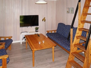 Sala de estar com televisão, mesa de centro de madeira, sofá azul estampado, escada de madeira e uma parede branca com linhas verticais.