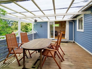 Eine überdachte Terrasse im Freien mit einem Holztisch und -stühlen, einer weißen Brüstung und Blick auf das Haus.