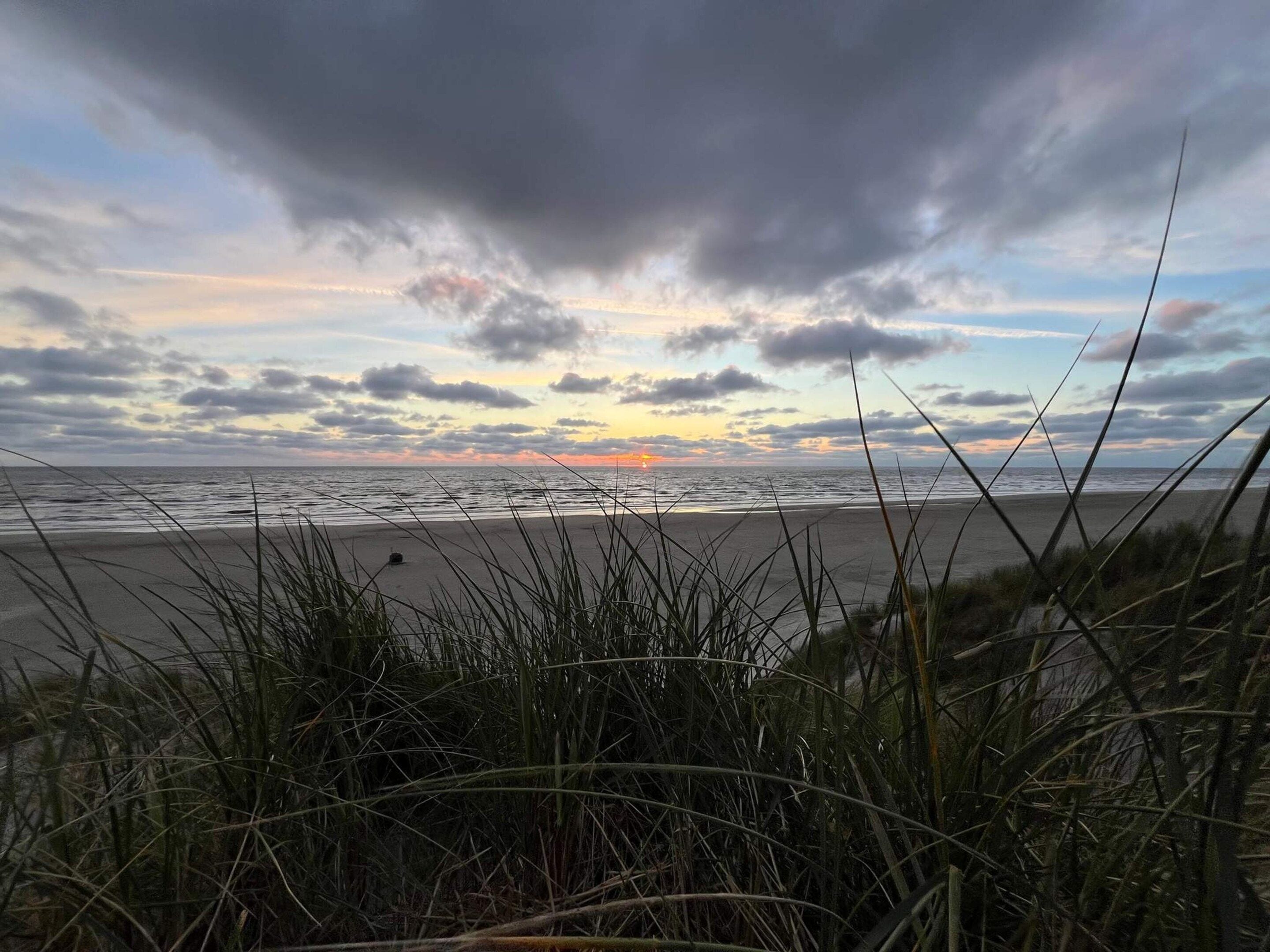 A beach with a sunset, grassy dunes, and a cloudy sky.