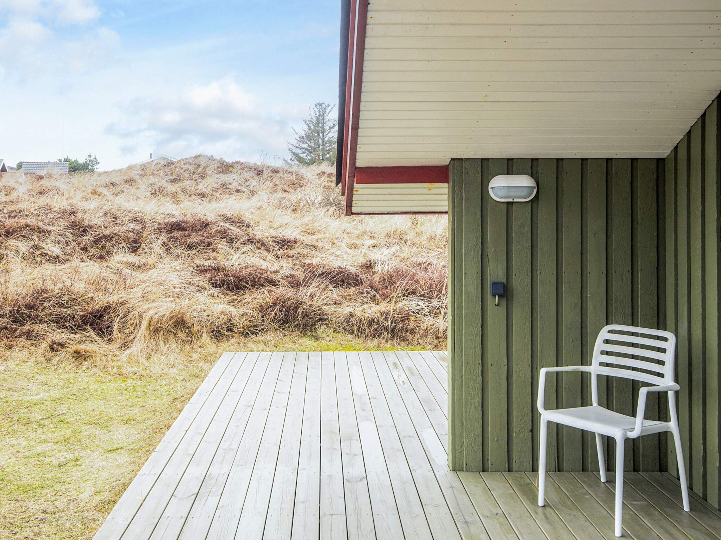 A wooden deck with a white plastic chair, a light fixture, and a view of a grassy hill.