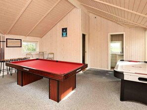 Mesa de bilhar com feltro vermelho, mesa de futebol de mesa e área de estar com cadeiras e mesa.