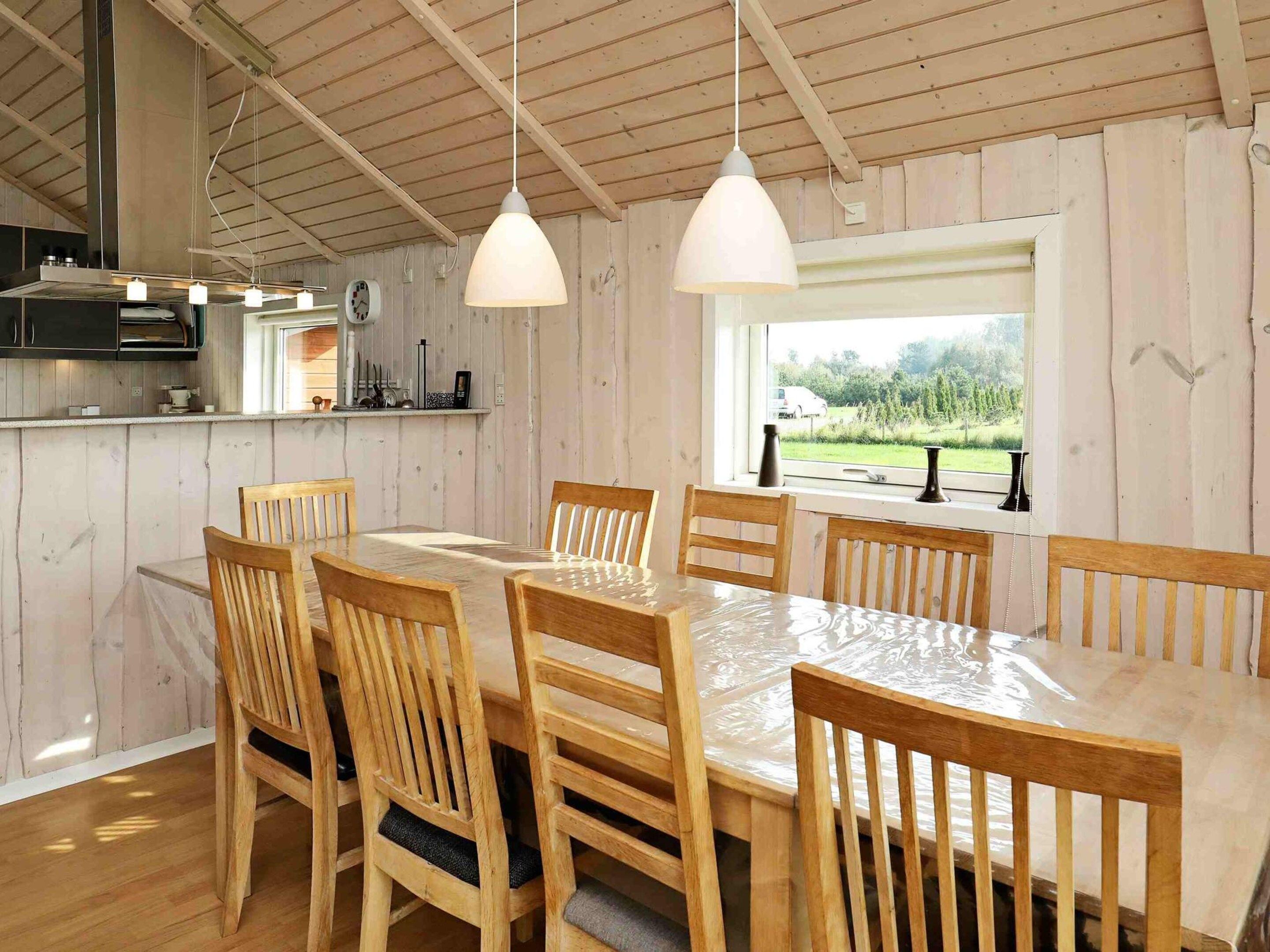 A wooden interior with a dining table and chairs, a kitchen area with cabinets and a clock, and a window with a view of greenery.