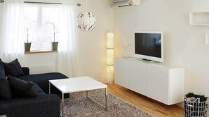 Una sala de estar moderna con un sofá oscuro, una mesa de centro blanca, un televisor de pantalla plana sobre un mueble blanco, una alfombra estampada y una planta en maceta cerca de la ventana.