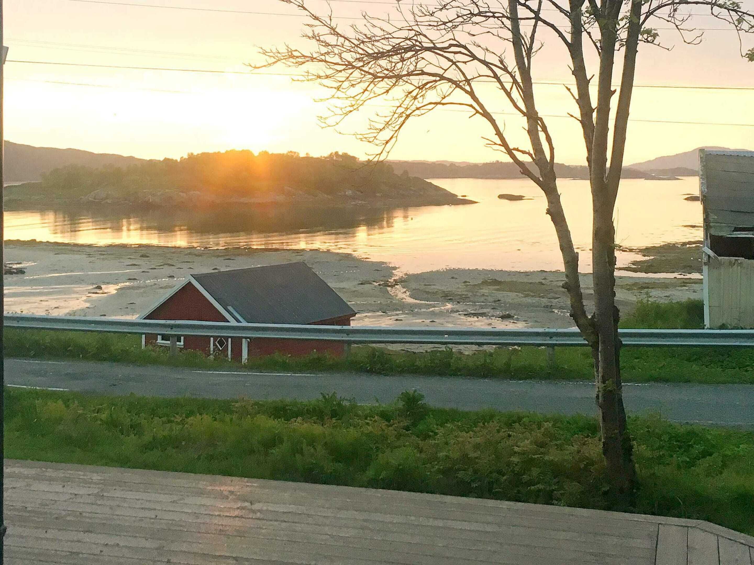 A coastal landscape with a small red building, a tree, and a body of water reflecting the sunset.