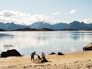 A beach with two people near rocks, a calm lake, and mountains in the background.