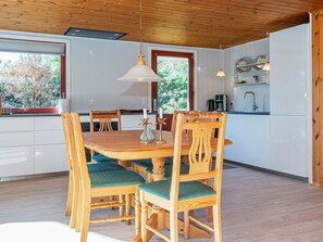 A wooden dining table with six chairs, a kitchen area with white cabinets, and a window with a view of trees. - 4 Person Holiday Home in Ulfborg (Ulfborg)