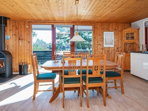 A wooden interior with a dining table, chairs, a fireplace, and a view through large windows.