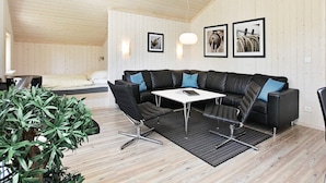 Ein modernes Wohnzimmer mit einem Ecksofa, einem Couchtisch und einem Esstisch. An der Wand hängen gerahmte Bilder und in der Ecke steht ein Blumentopf.