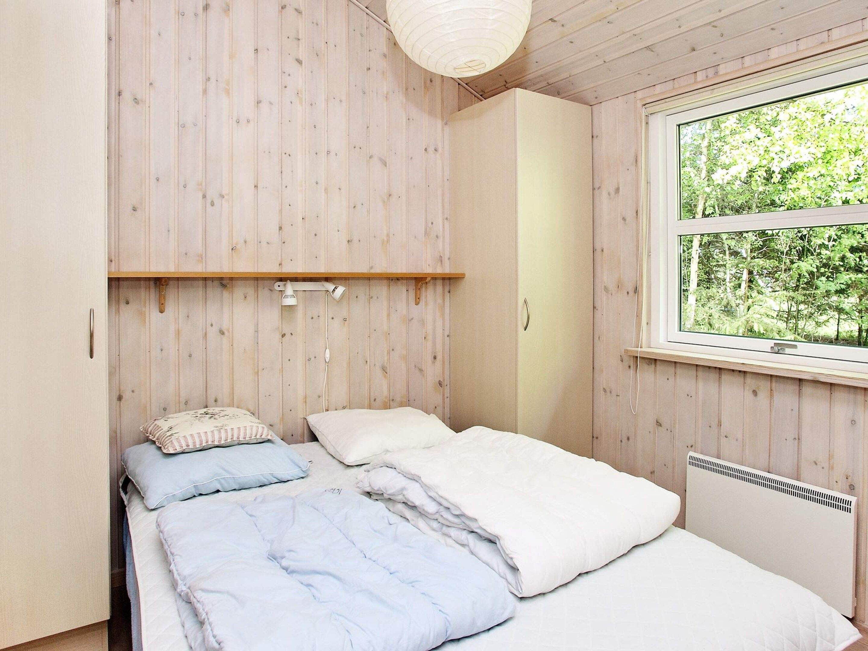 Ein Schlafzimmer mit einem Bett, einem Nachttisch, einer Lampe und einem Fenster mit Blick auf Bäume.