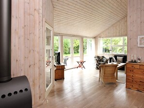 A room with a large fireplace, a television, wicker furniture, and a view of greenery through large windows.