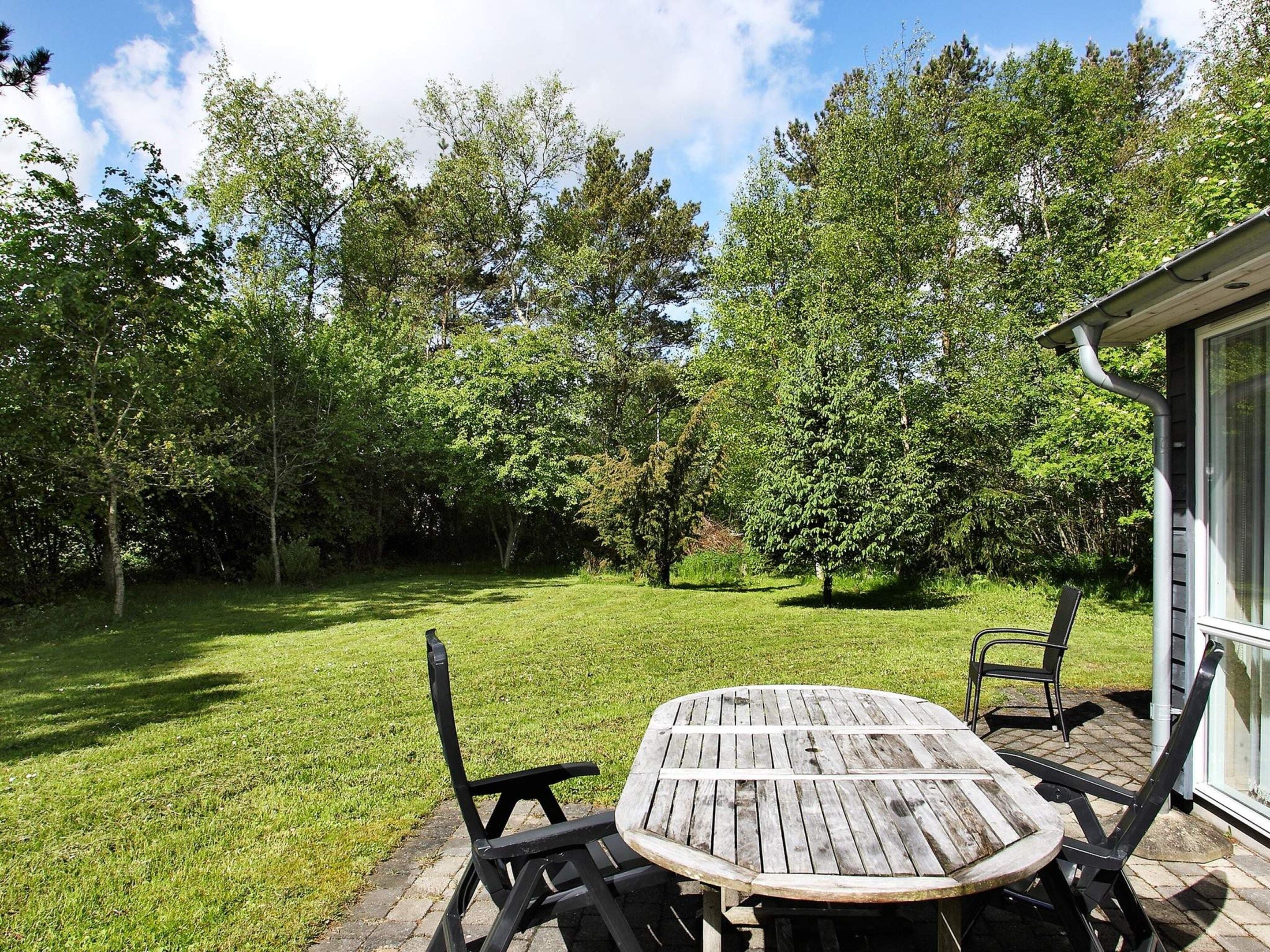 A patio area with a round wooden table and two chairs, surrounded by a grassy lawn and trees.