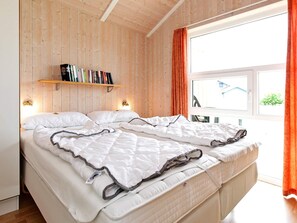 Cama doble con ropa blanca, pared de madera, estante con libros y ventana con cortinas.