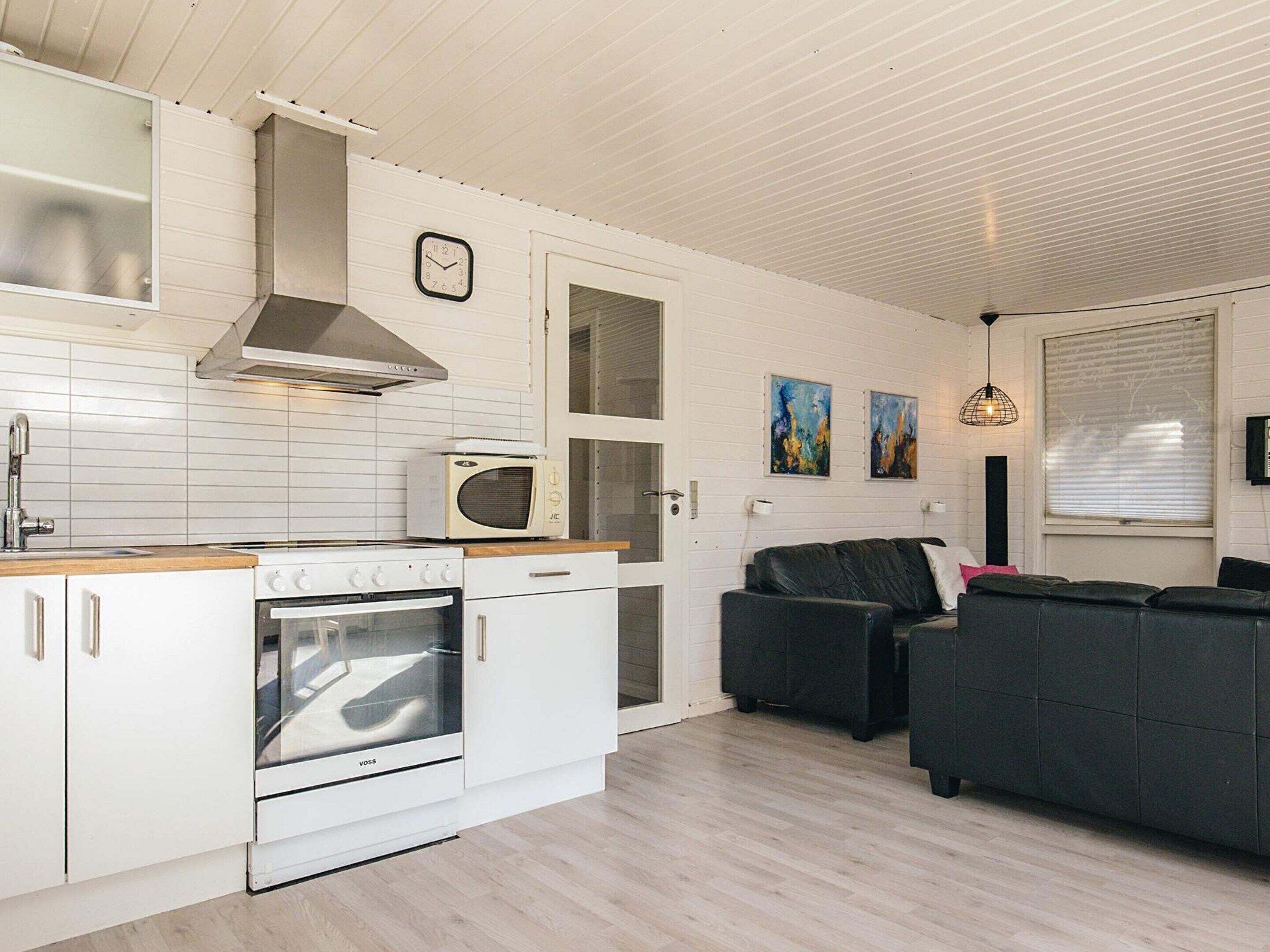 A compact kitchen with white cabinetry, a built-in oven, and a microwave. There is a dining area with a black sofa and a window with blinds.