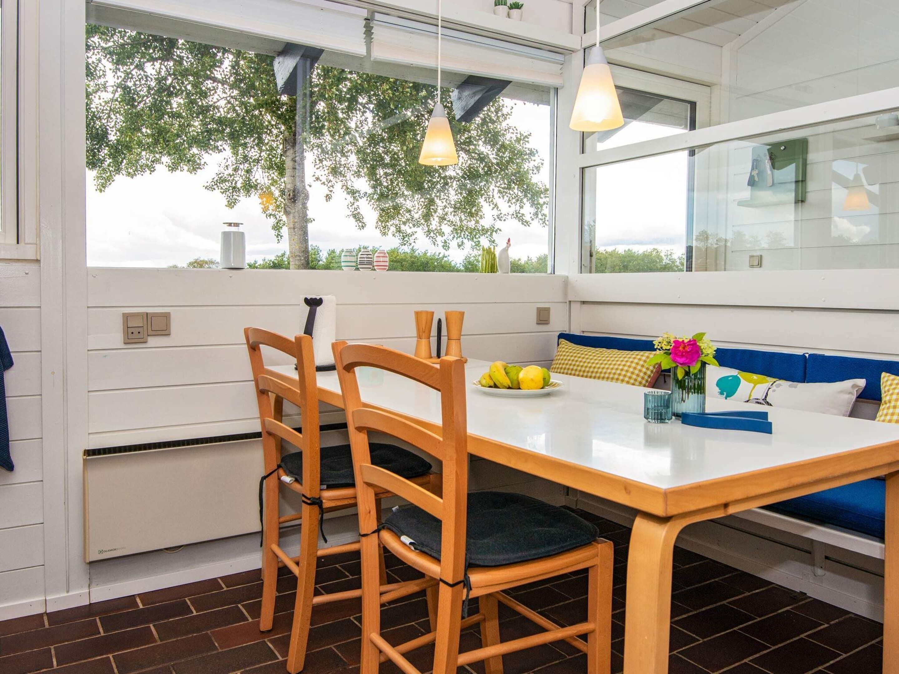 A modern kitchen with a wooden table and chairs, a built-in bench, and large windows offering a view of trees.