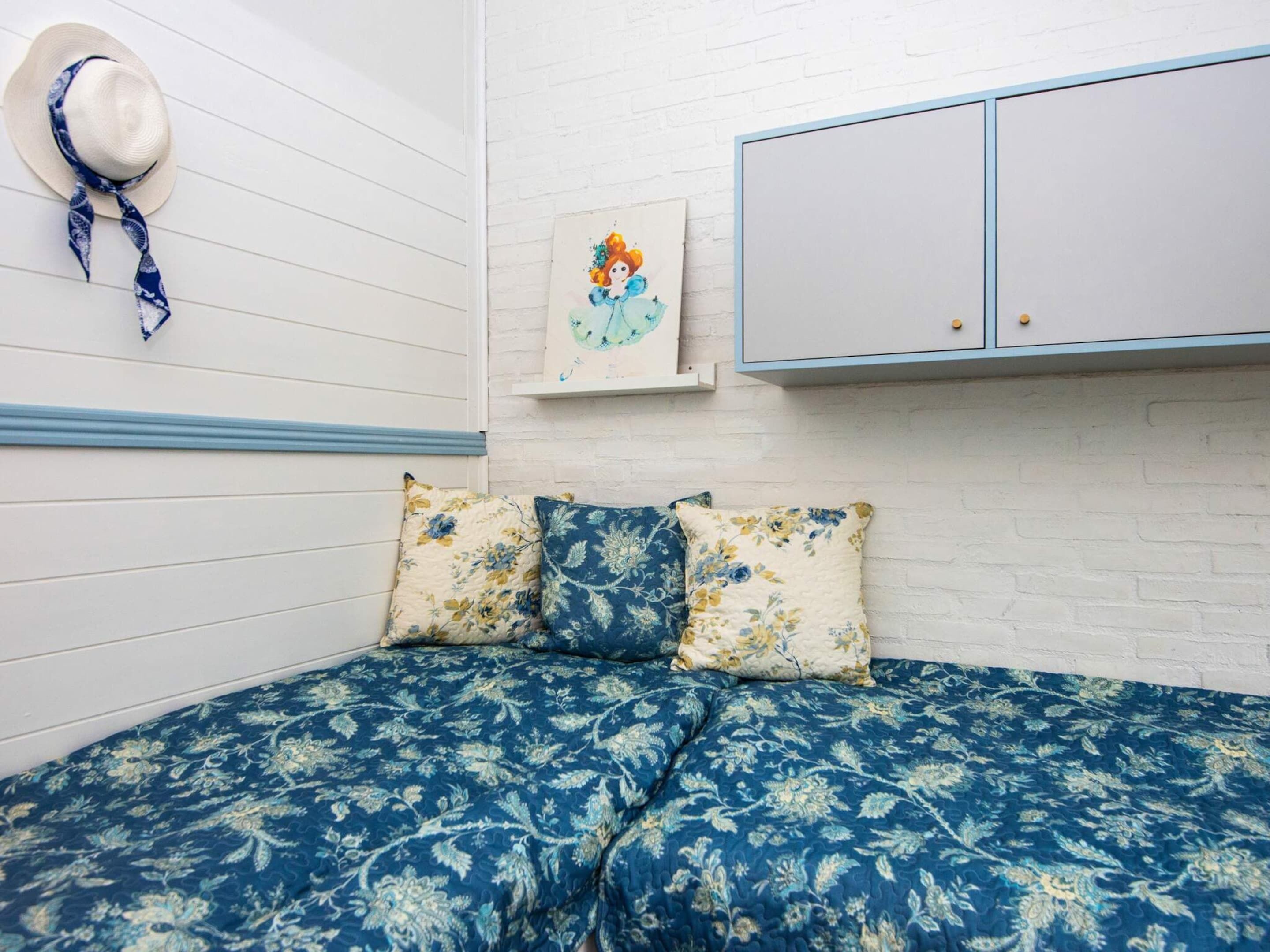 A bedroom with a bed, floral pillows, a hat, and a painting on the wall.